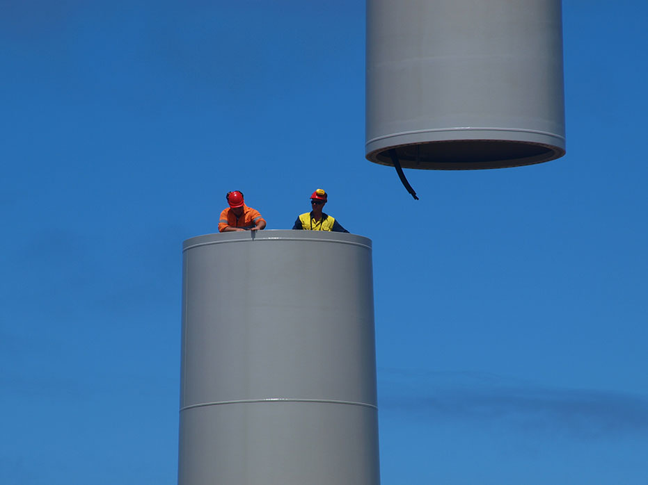 A photo of 2 people with their upper torsos protruding from one of the upright tower sections, preparing to assemble the next tower section as it is lowered into place.