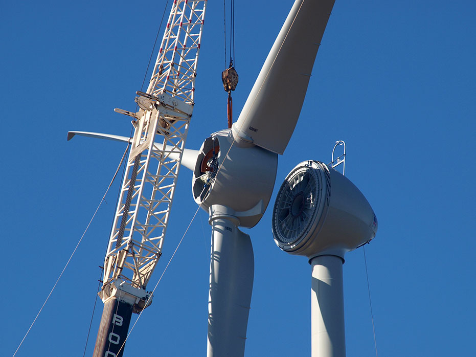 A photo of the hub and blades assembled and about to be placed into position on the nacelle by a crane.